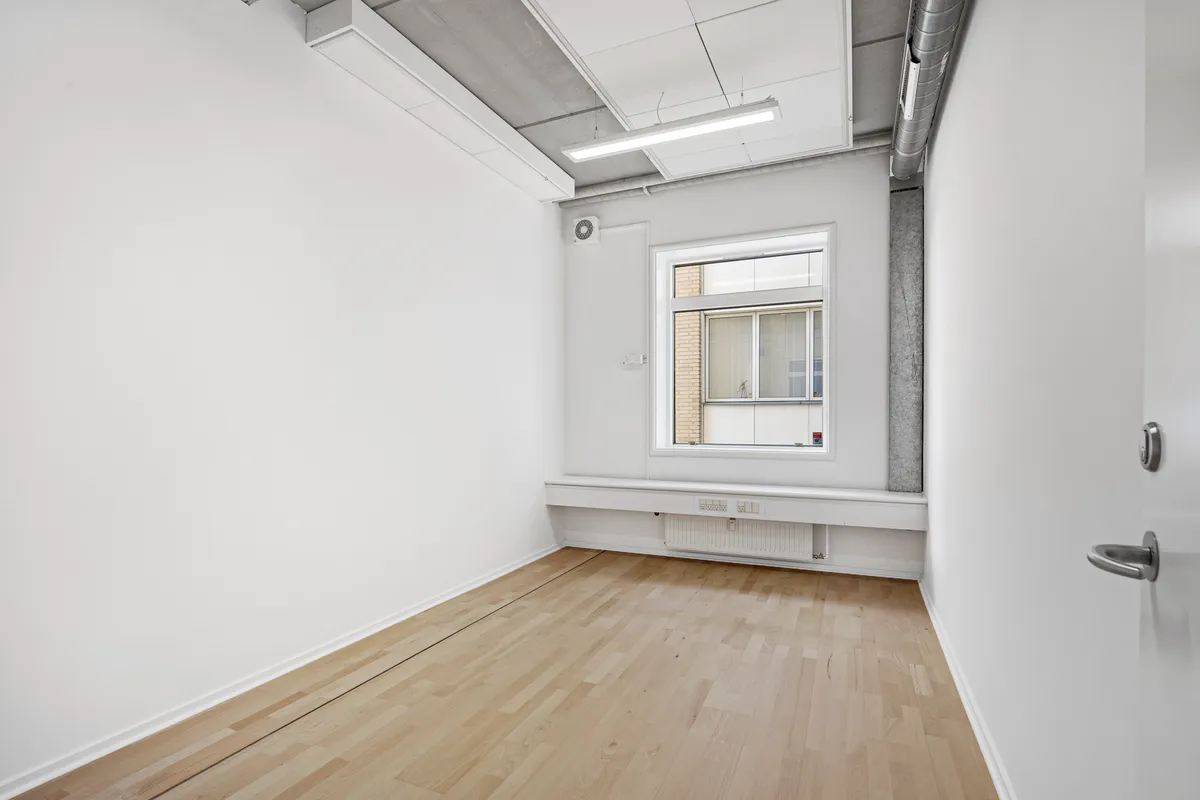 Et tomt kontor med lyse hvide vægge og gulvtæppe, vindueskarmen med radiator, og lyskilder i loftet. Der er en håndtag på højre side af døren og en hvid ventilationsblok på væggen