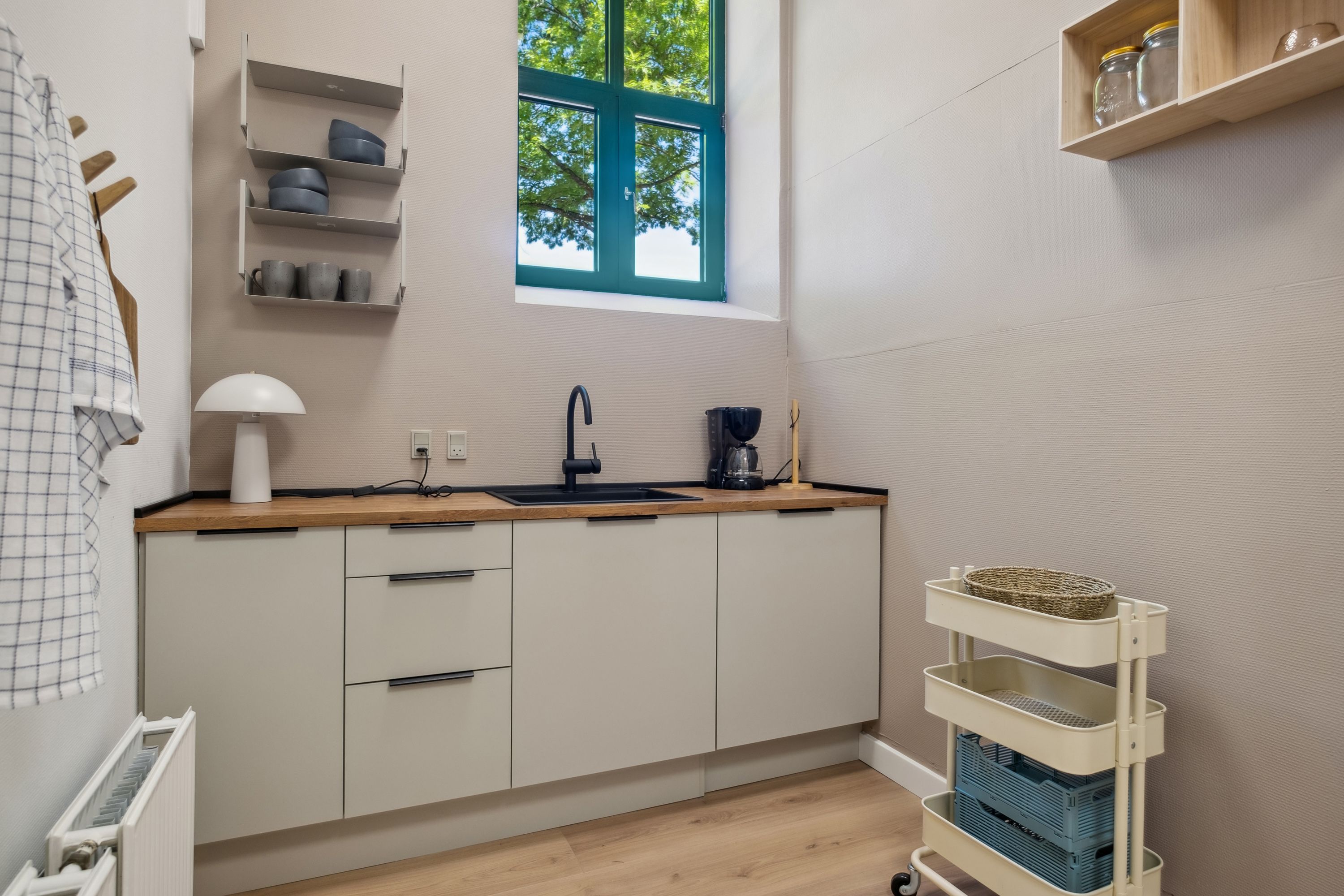 En lille køkken med hvide vægge, lyse trægulve og hvide skabe. På en høj hylde står der glas og kopper. Et skarpt vindue med