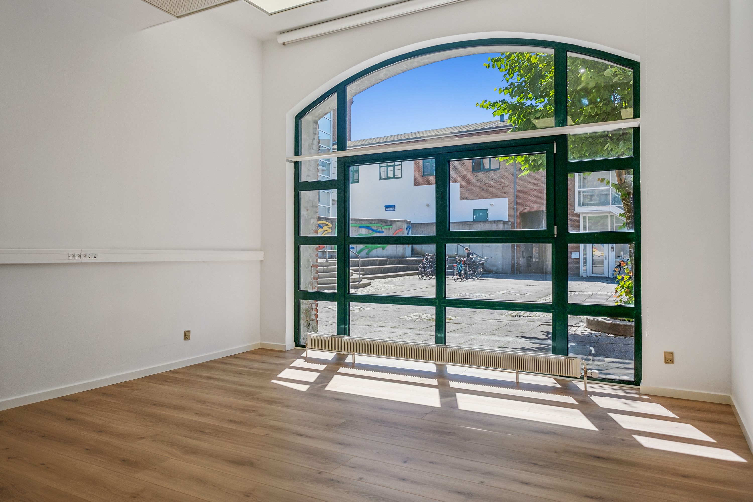 Lokal med trægulv og hvide vægge samt glasdør med udsigt til gågade med både skulpturer og cykler samt bygning med facade