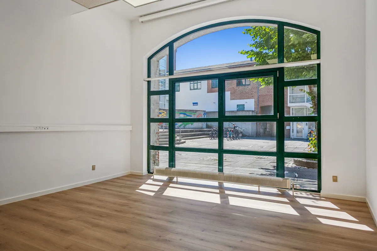 Lokal med trægulv og hvide vægge samt glasdør med udsigt til gågade med både skulpturer og cykler samt bygning med facade