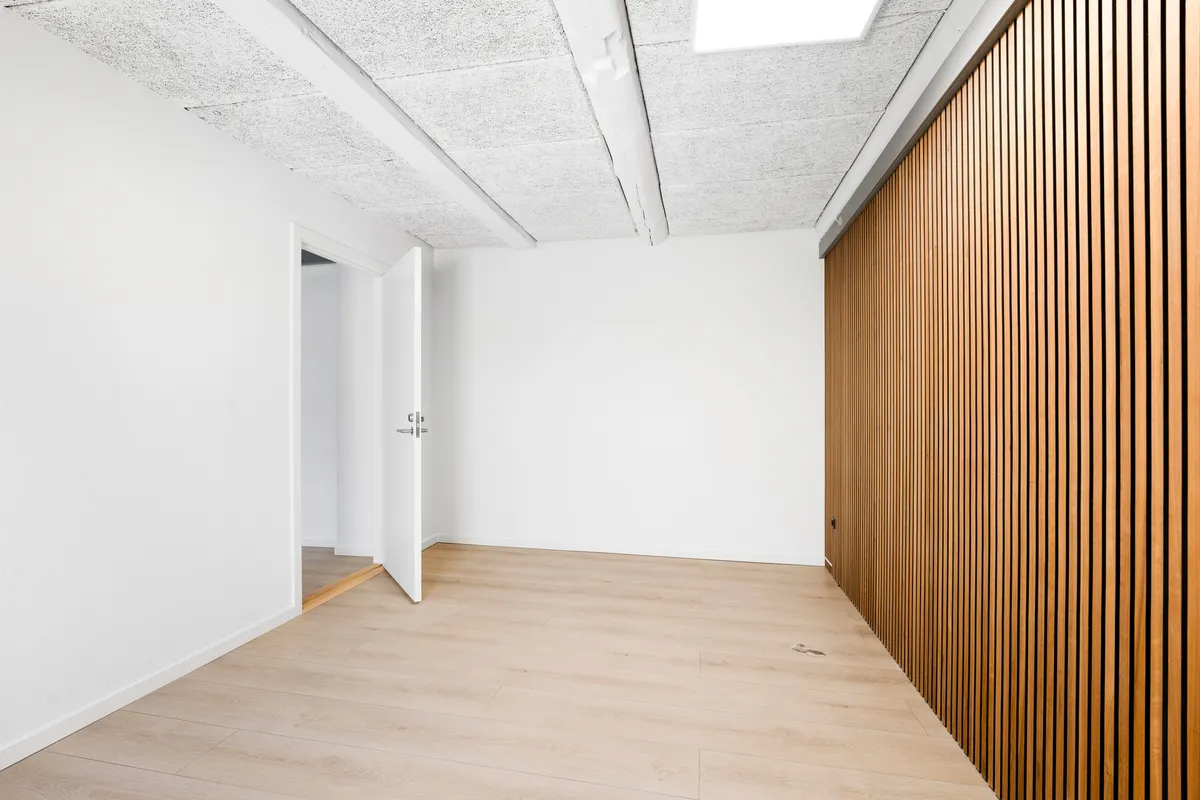 En minimalistisk, ubeboet og tæt rum med hvide vægge, lyse gulvbelægning og en åben dør med en smal gang til et andet rum