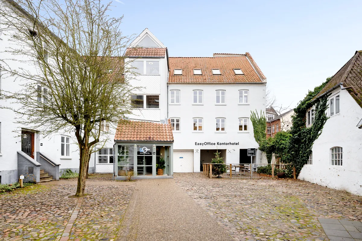 Hjørnet af et hus med navnet EasyOffice Kontorhotel, gårdsplads med en grøn plante i potte og et bænk på den anden side af