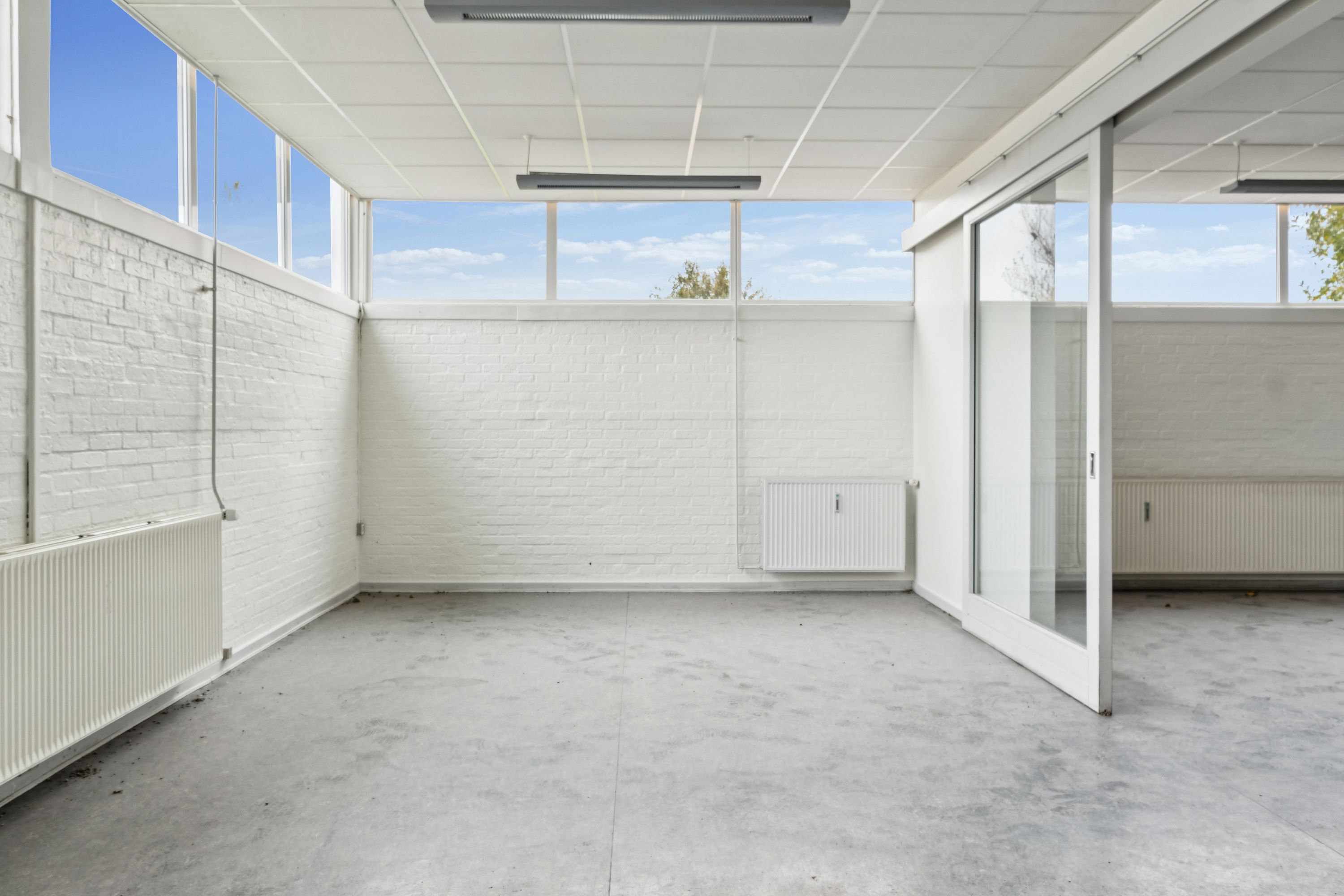 Et tomt rum med hvide vægge og loft, store vinduer, glasdøre og radiatorer samt to lysekroner og et højt loft med lyskryds