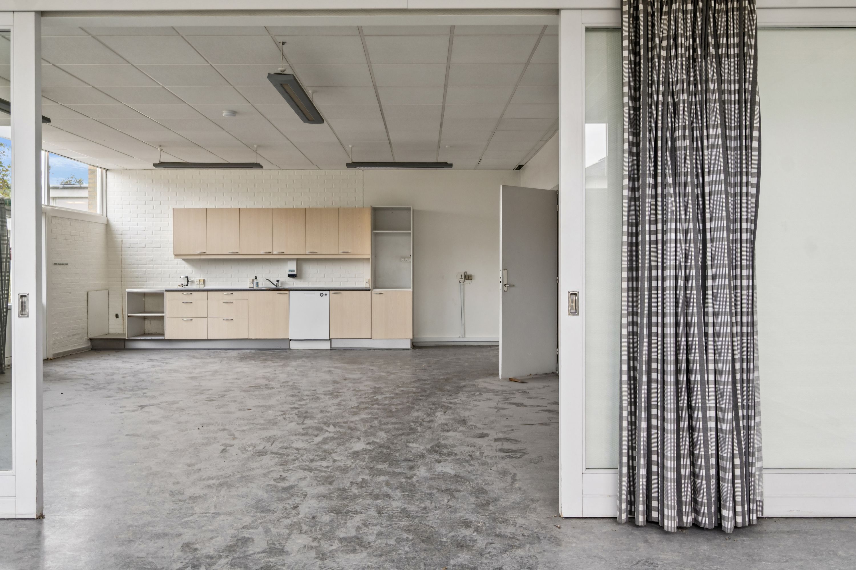 Et tomt lager med køkken og åbne døre, hvor en delvis åben dør viser ud til et tomt gulv med belysning fra loftet