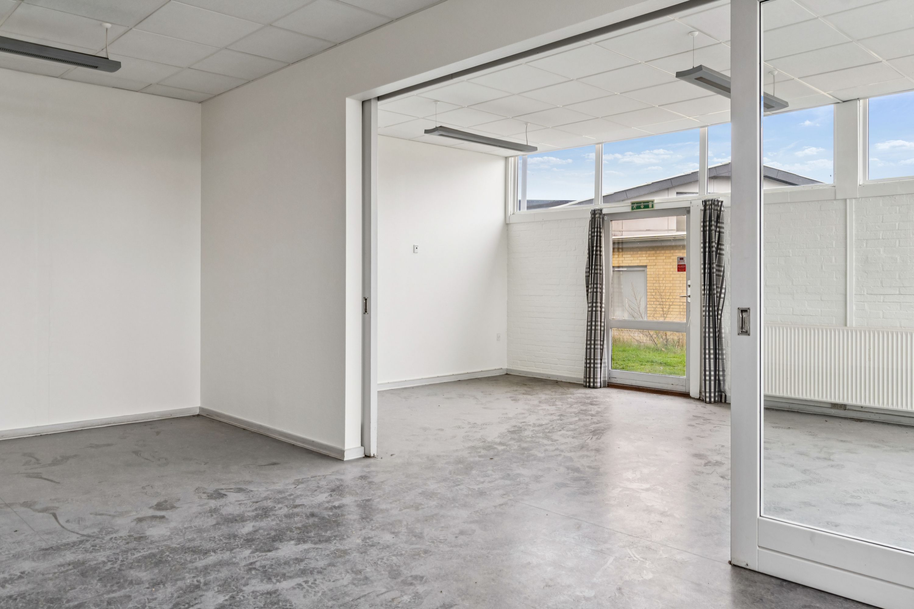 En tom og ren lejlighed med hvide vægge og cementgulv og store vinduer med udgang til en veranda med græsplæne og bygning i