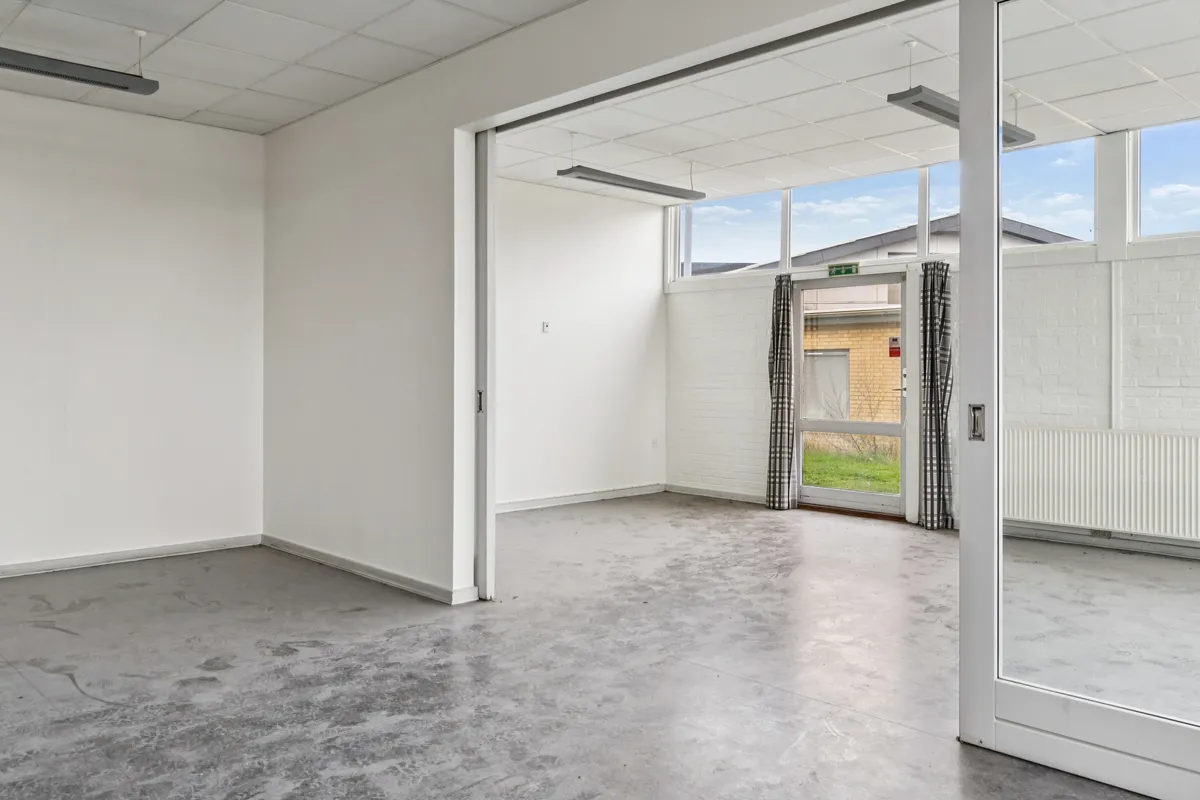 En tom og ren lejlighed med hvide vægge og cementgulv og store vinduer med udgang til en veranda med græsplæne og bygning i