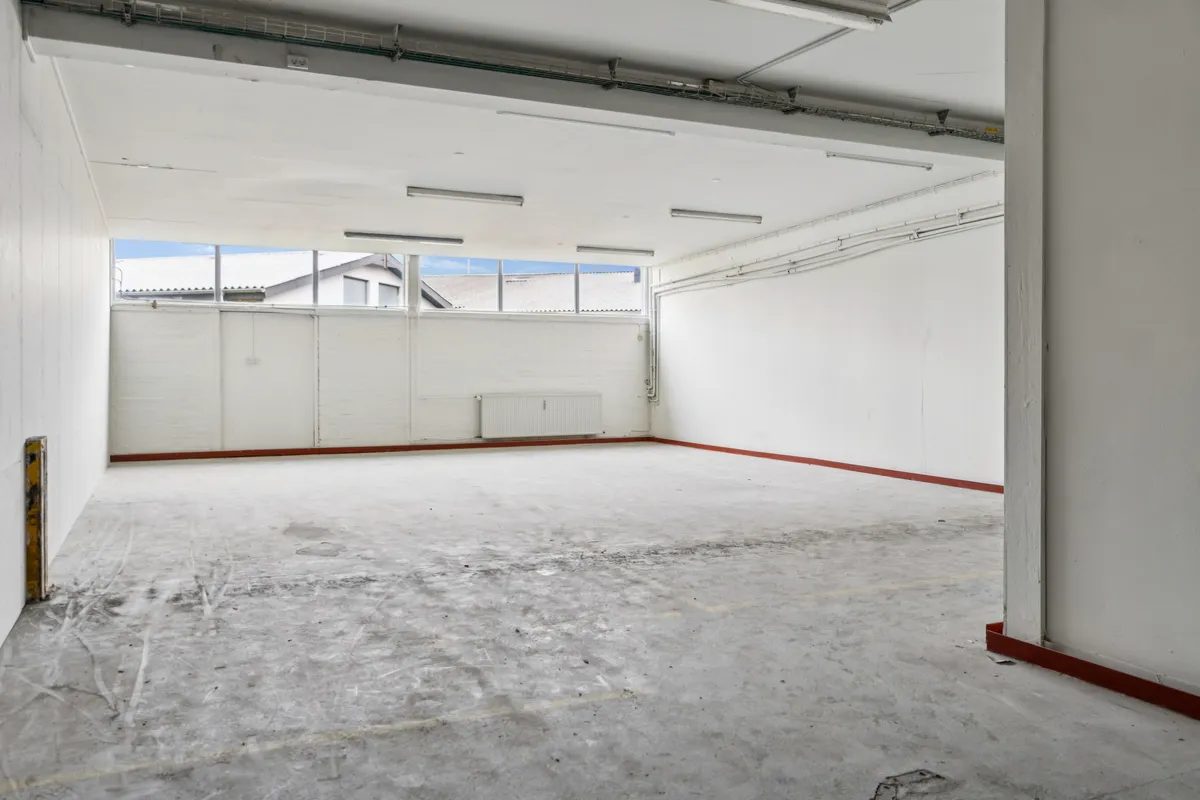 Et stort tomt lager med hvide vægge og cementgulv, vinduer mod gårdspladsen og røde kantbånd langs gulvet
