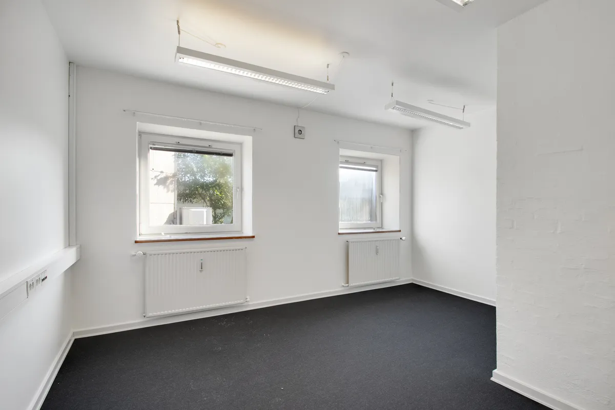 Et tomt rum med hvide vægge og gulv samt to vinduer med radiatorer og lysekroner