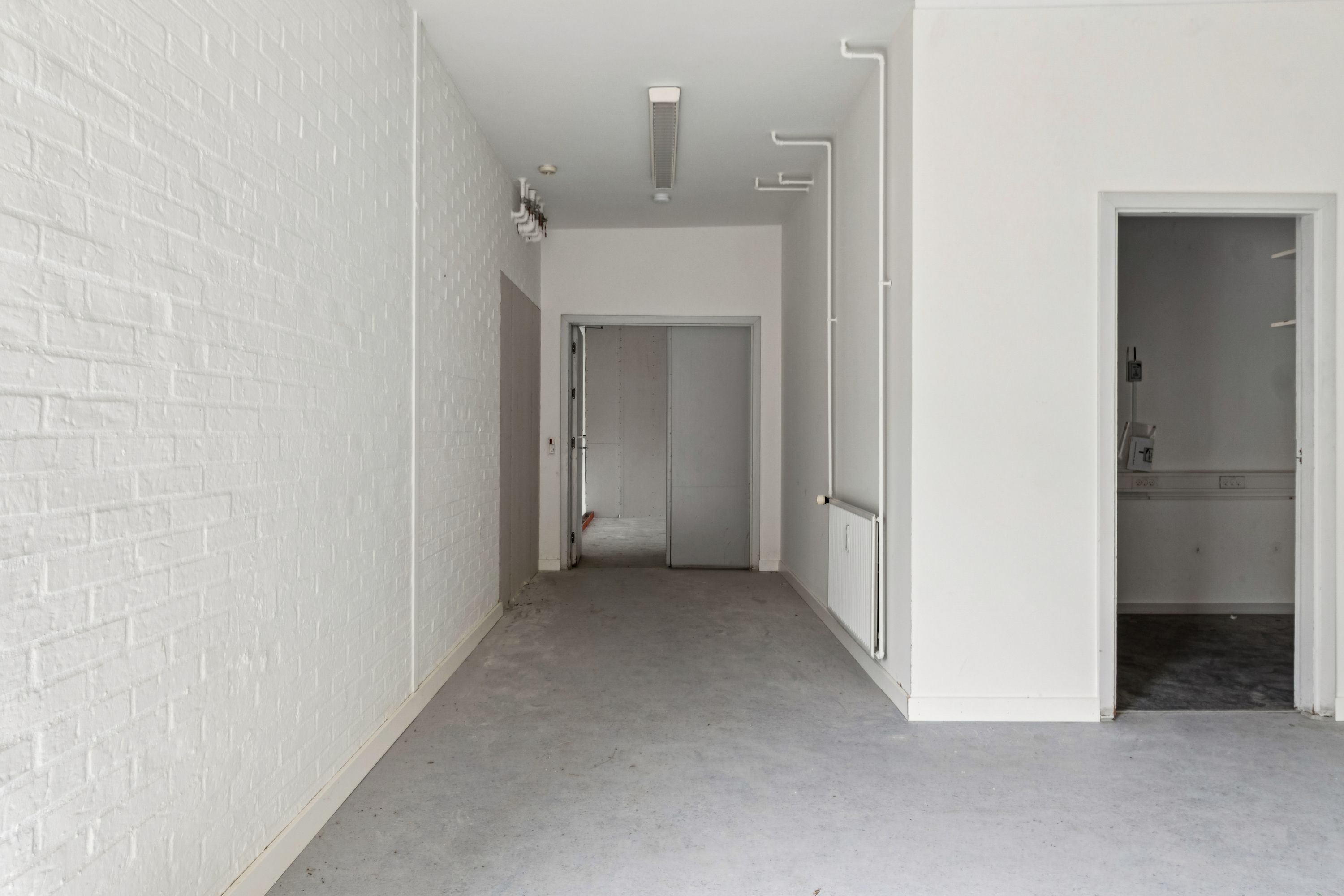 En lang og smal gang med hvide vægge og lyse gulv, to døre og et skab til højre samt en hvid radiator og en hvid loftslampe