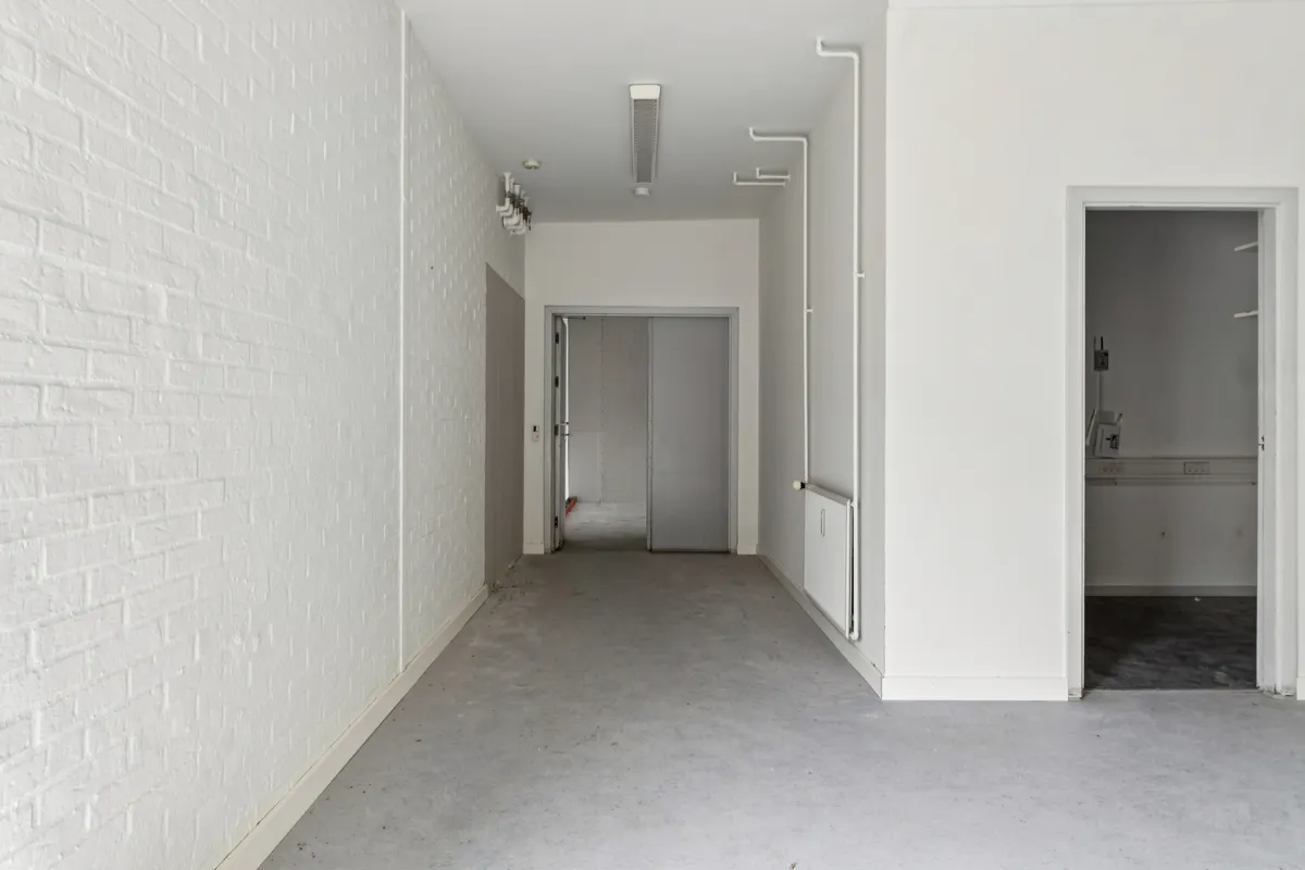 En lang og smal gang med hvide vægge og lyse gulv, to døre og et skab til højre samt en hvid radiator og en hvid loftslampe