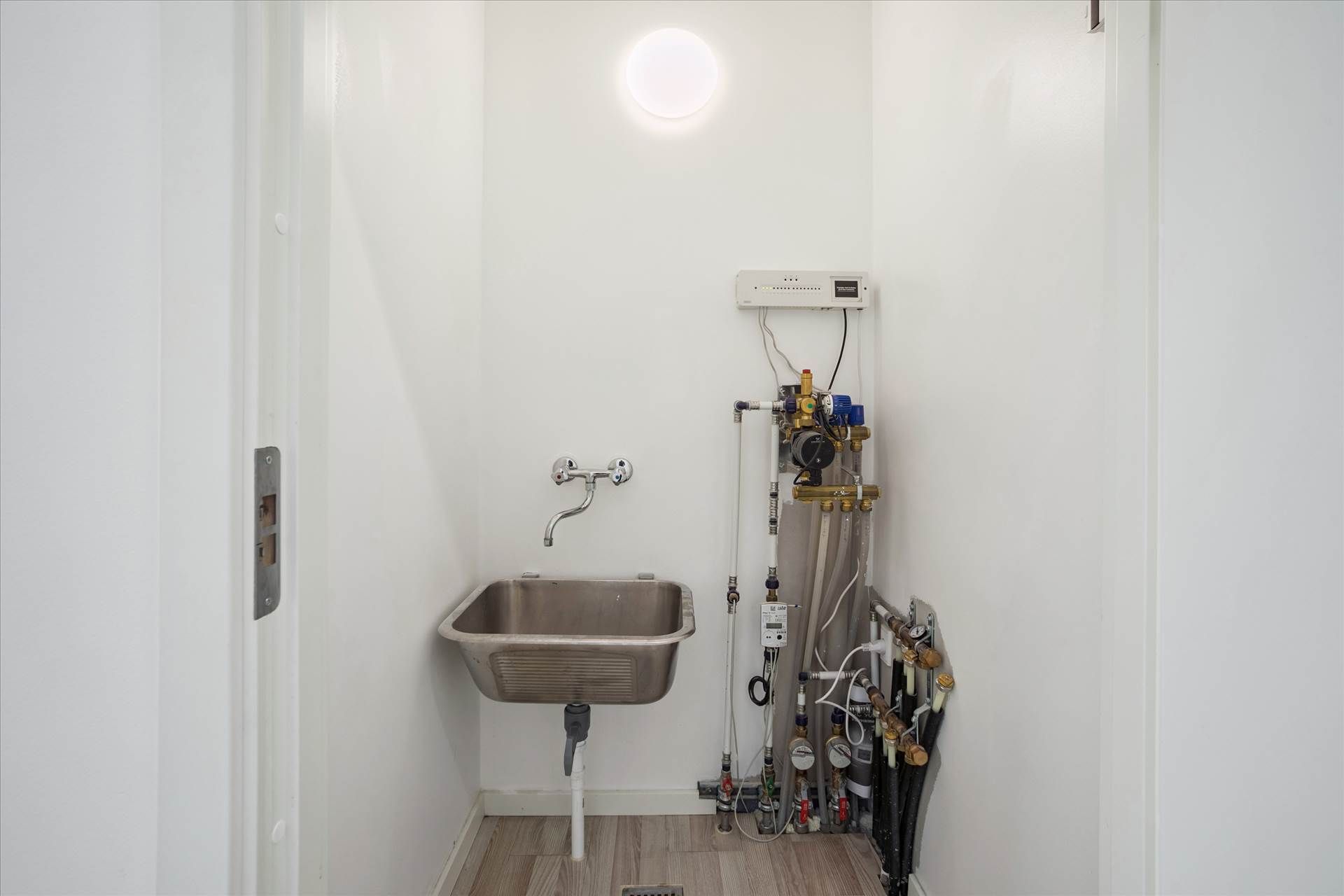 Værelse med hvid væg, håndvask, lys, varmepumpe, manometer og andre rørværk samt en gulvdrain.
