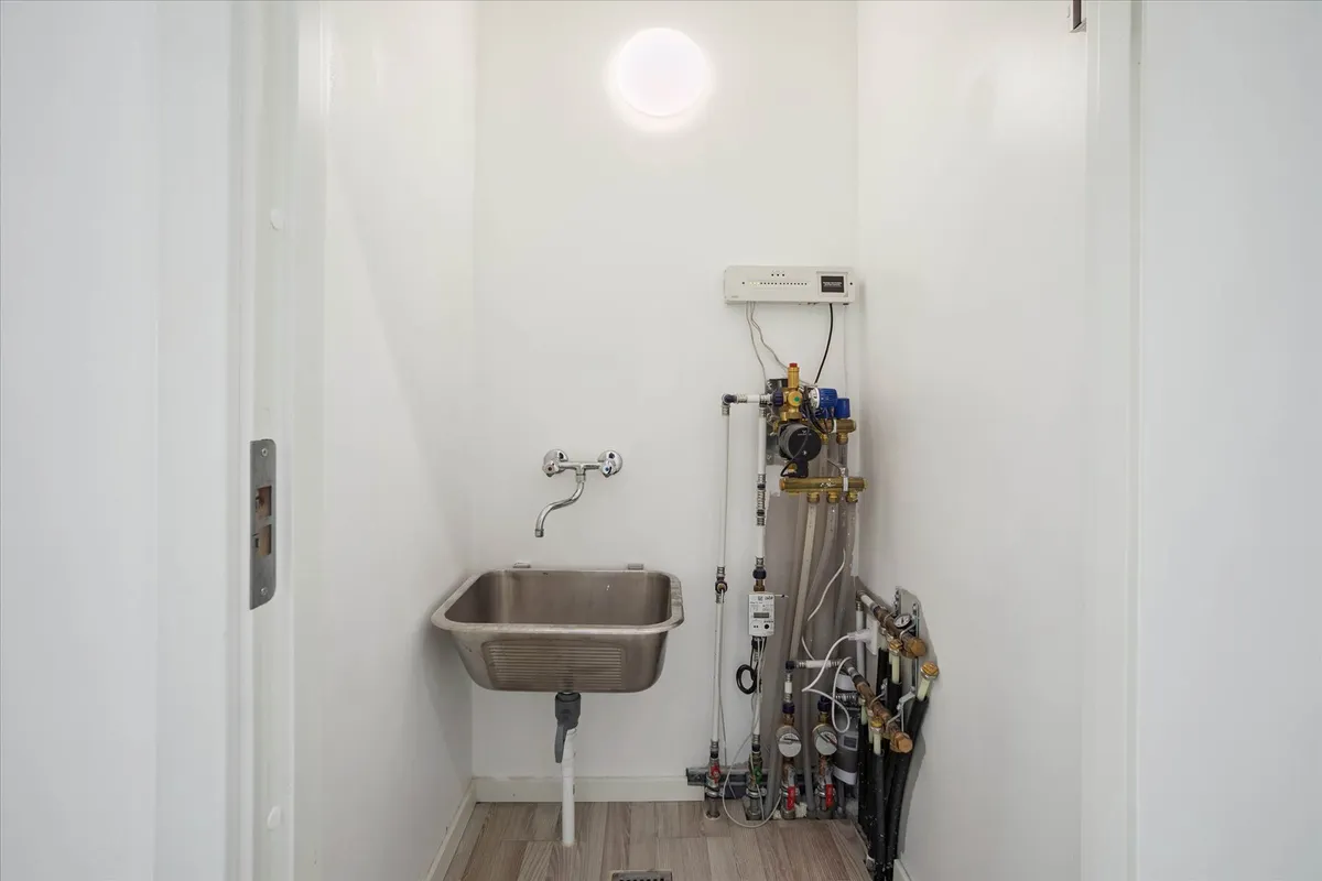 Værelse med hvid væg, håndvask, lys, varmepumpe, manometer og andre rørværk samt en gulvdrain.
