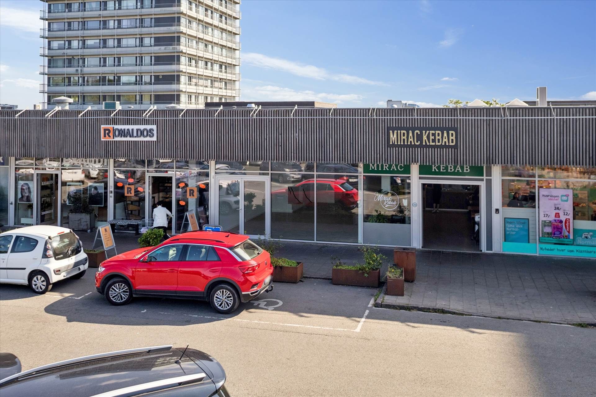 Forretningslokaler med butikker, Mirac Kebab og Ronalds, med biler parkeret på fortovet og i garagen, baggrund med lejemål