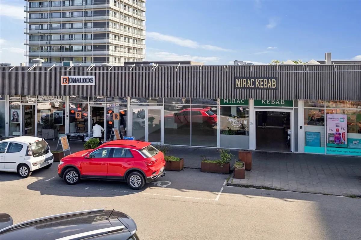 Forretningslokaler med butikker, Mirac Kebab og Ronalds, med biler parkeret på fortovet og i garagen, baggrund med lejemål