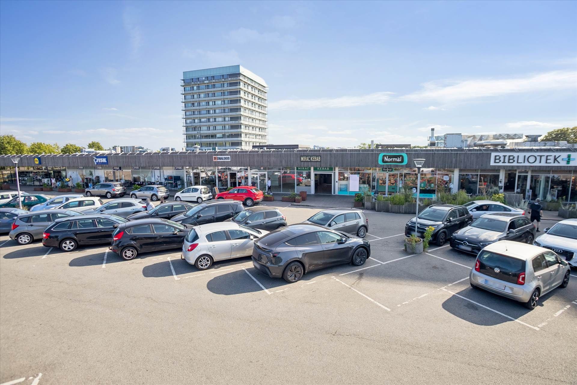 Parkeringsplads foran diverse butikker som Normal, Mirak Kebab og Bibliotek med biler parkeret og gående mennesker