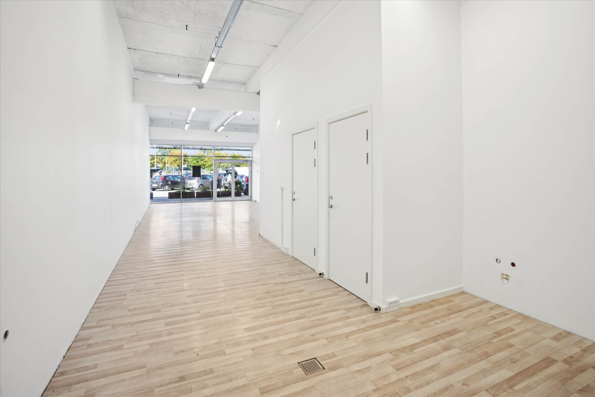 Store rum med hvide vægge og lysegult trægulv og glasdøre med vinduer ude på parkeringspladsen med biler og planter i