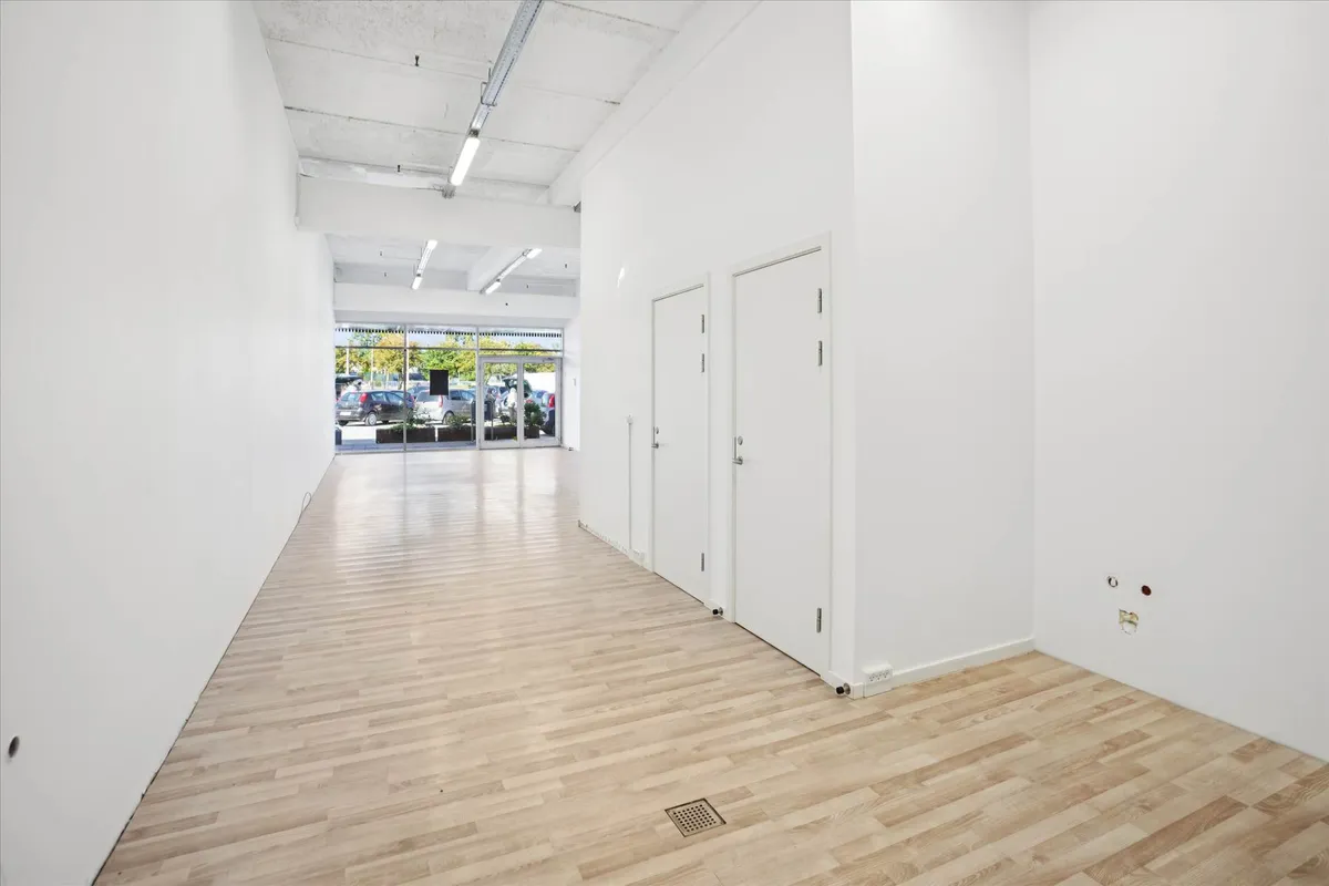 Store rum med hvide vægge og lysegult trægulv og glasdøre med vinduer ude på parkeringspladsen med biler og planter i