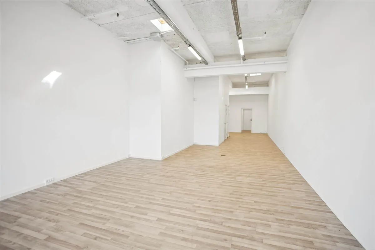 Lejlighed med lyse trægulve og hvide vægge, lyse gulvlampe og et lille vindueslys, der falder på gulvet. Lejligheden er tæt