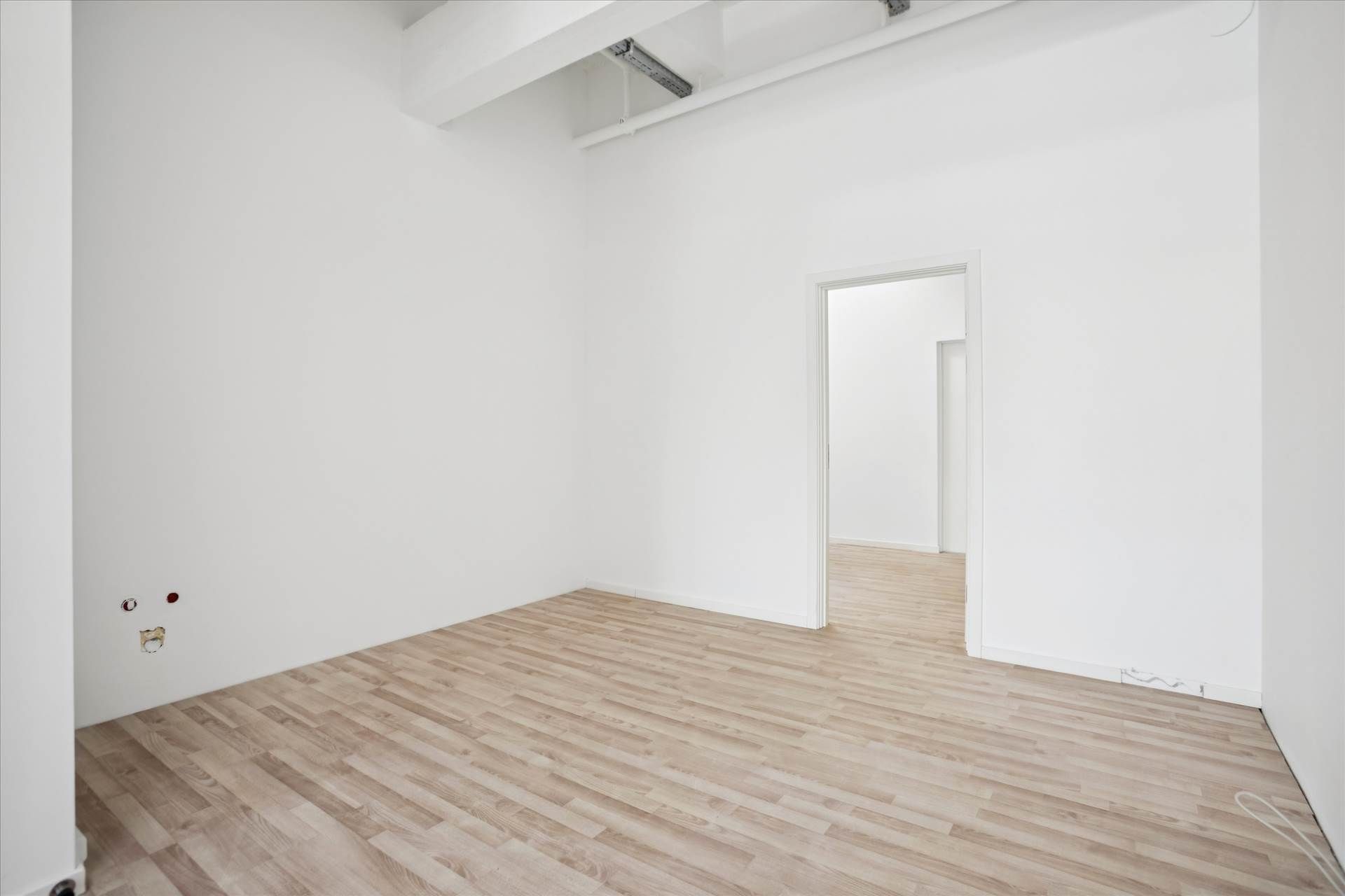 Tomt rum med hvide vægge og lyse trægulve. Et hvidt dørkarm med åben dør. Væggen er ubehandlet og har hul efter stikkontakt.