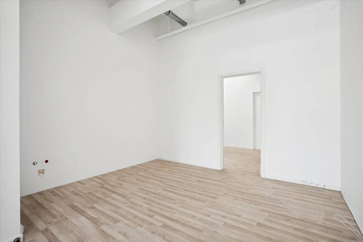 Tomt rum med hvide vægge og lyse trægulve. Et hvidt dørkarm med åben dør. Væggen er ubehandlet og har hul efter stikkontakt.