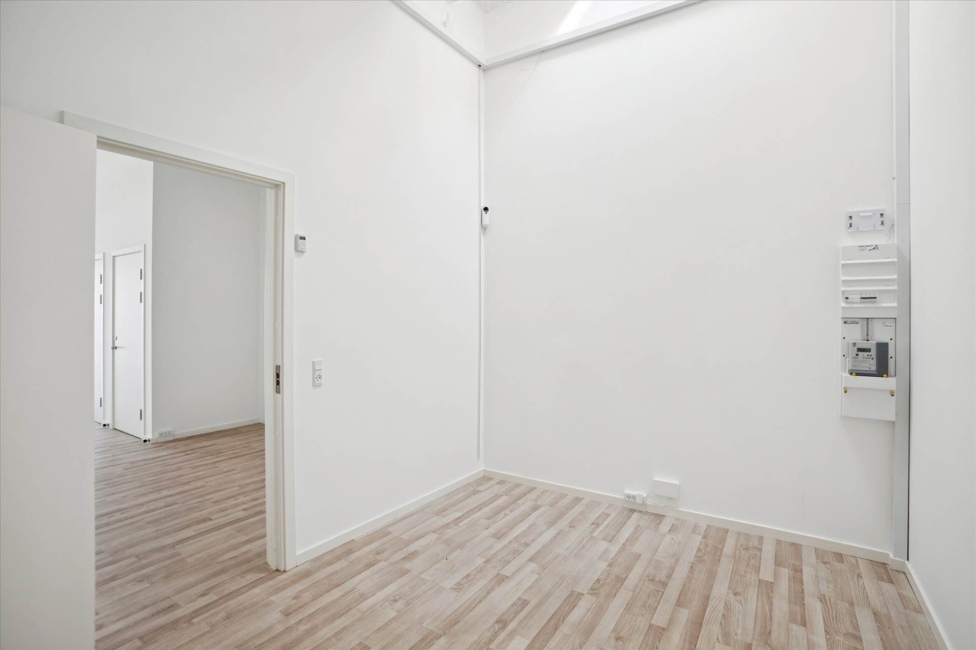 En tom stue med hvide vægge og gulvtæppe, der vender mod åbne døre til en anden rum. En hvid dør og et stikkontakt er