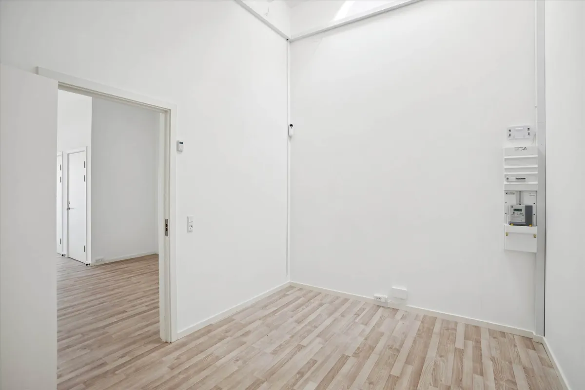 En tom stue med hvide vægge og gulvtæppe, der vender mod åbne døre til en anden rum. En hvid dør og et stikkontakt er