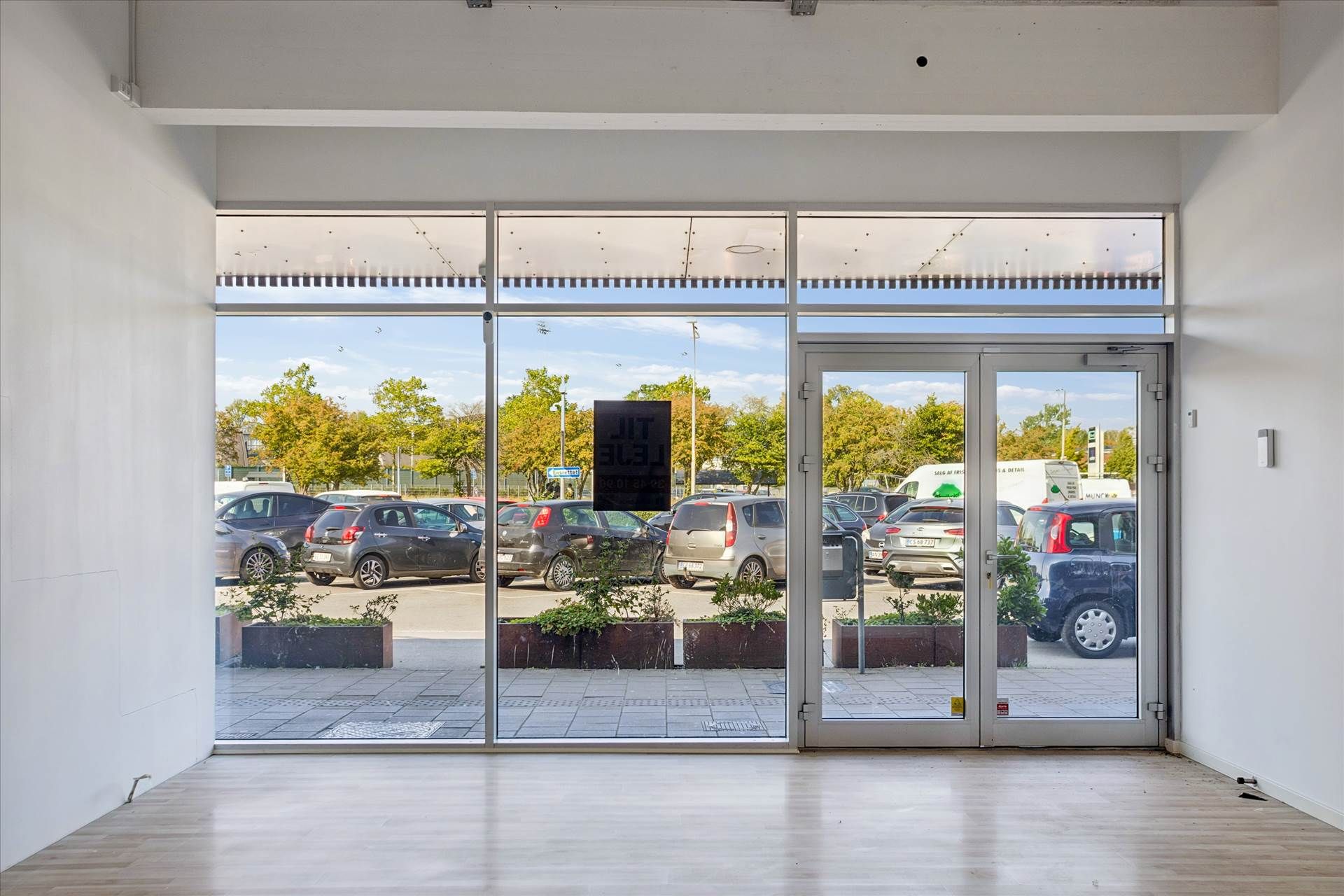Et forretningslejemål med glasdøre og plantepotter i en parkeringsplads med biler og træer