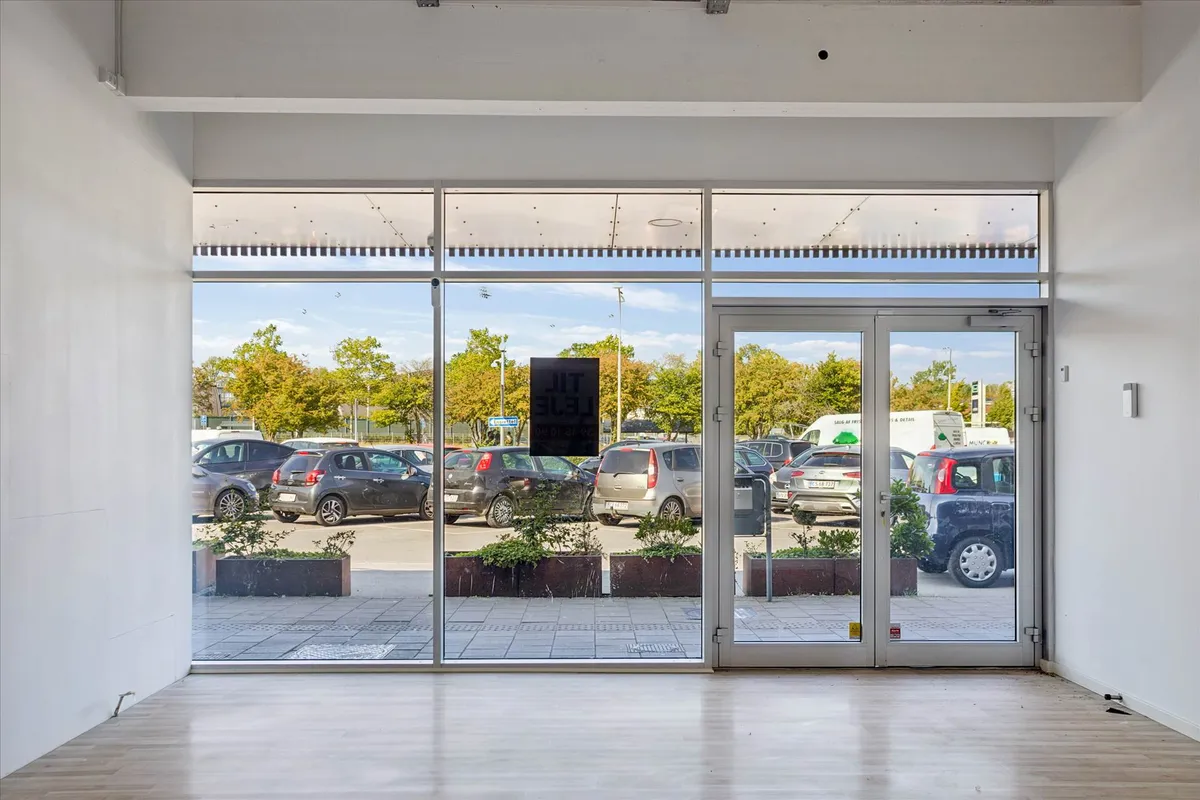 Et forretningslejemål med glasdøre og plantepotter i en parkeringsplads med biler og træer