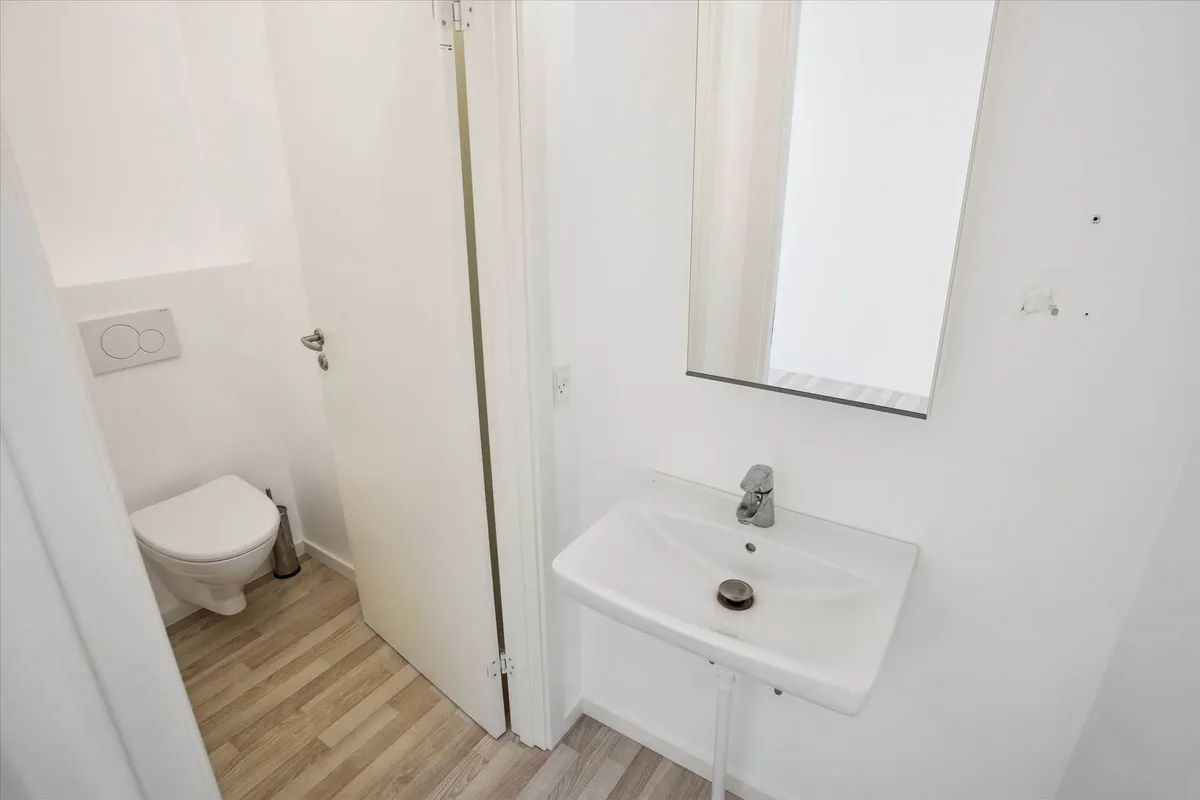 Lejlighed med hvidt toilet og hvid vask samt spejl og stikkontakt i hvid væg og gulvtæppe