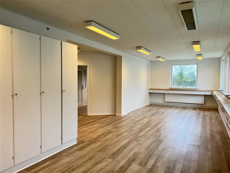 Et tomt kontor med hvide vægge, lysegul gulvbelægning, loftslampe, høj vinduesglas, åbne hvide kabinetter og et skrivebord