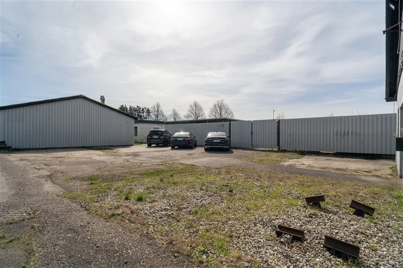 Tre biler parkeret foran en hvid mur. Området er omgivet af en høj metalgitter. Den lyse himmel er fyld af skyer.