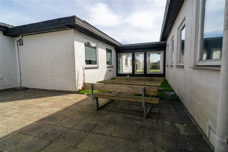 En moderne hvid bygning med en terrasse, der har et træbord og en bænk samt glasdøre