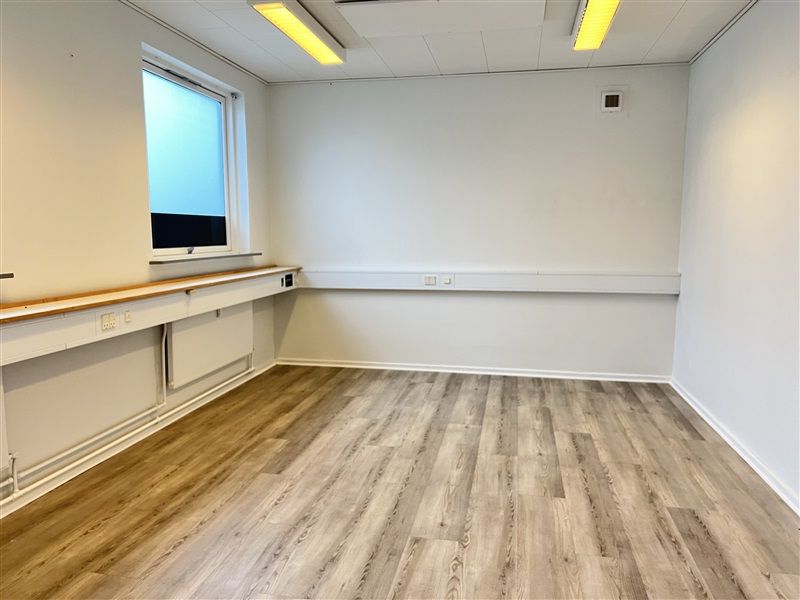 Kontorrum med lyse gulvbelægning og hvide vægge, to vinduer, hvide og brune skralde, samt to stikkontakter på væggen