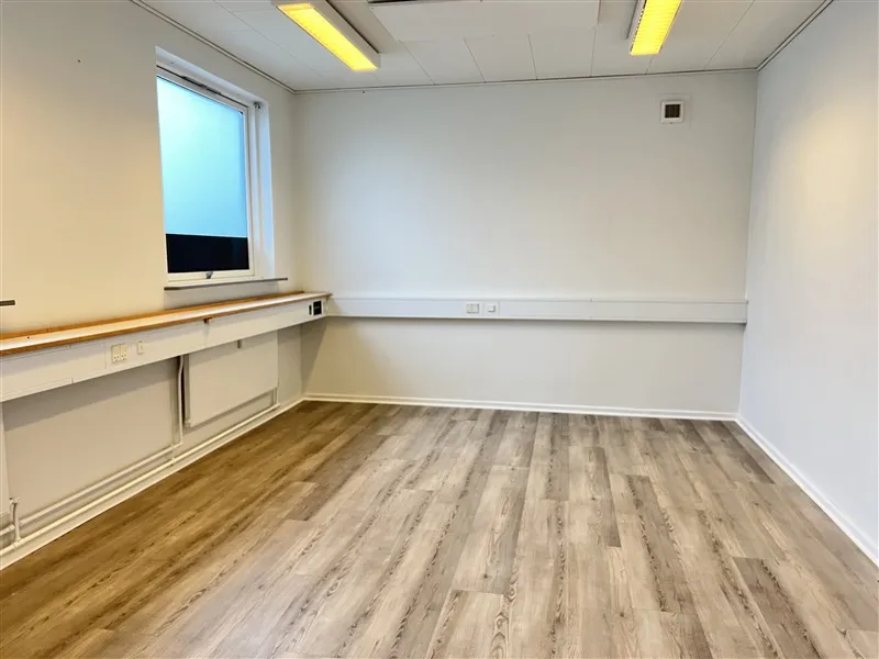 Kontorrum med lyse gulvbelægning og hvide vægge, to vinduer, hvide og brune skralde, samt to stikkontakter på væggen