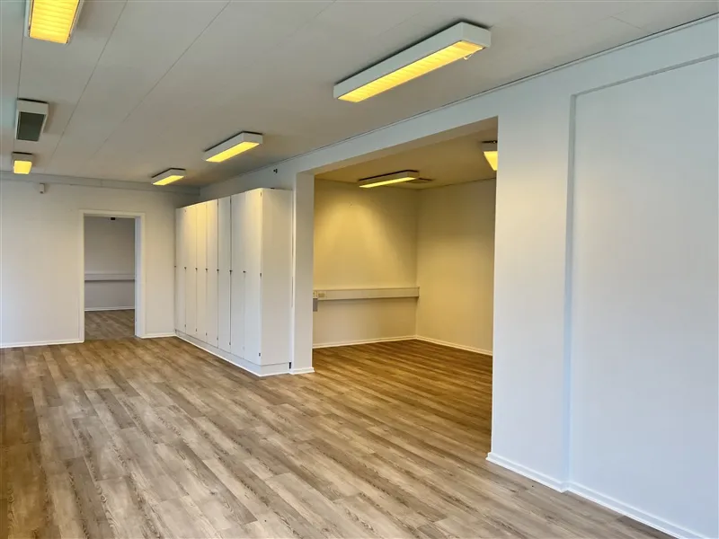 Moderne kontorlokaler med lyse loftslamper, hvide vægge og trægulv. På den ene side er der høje hvide skabe, og på den anden