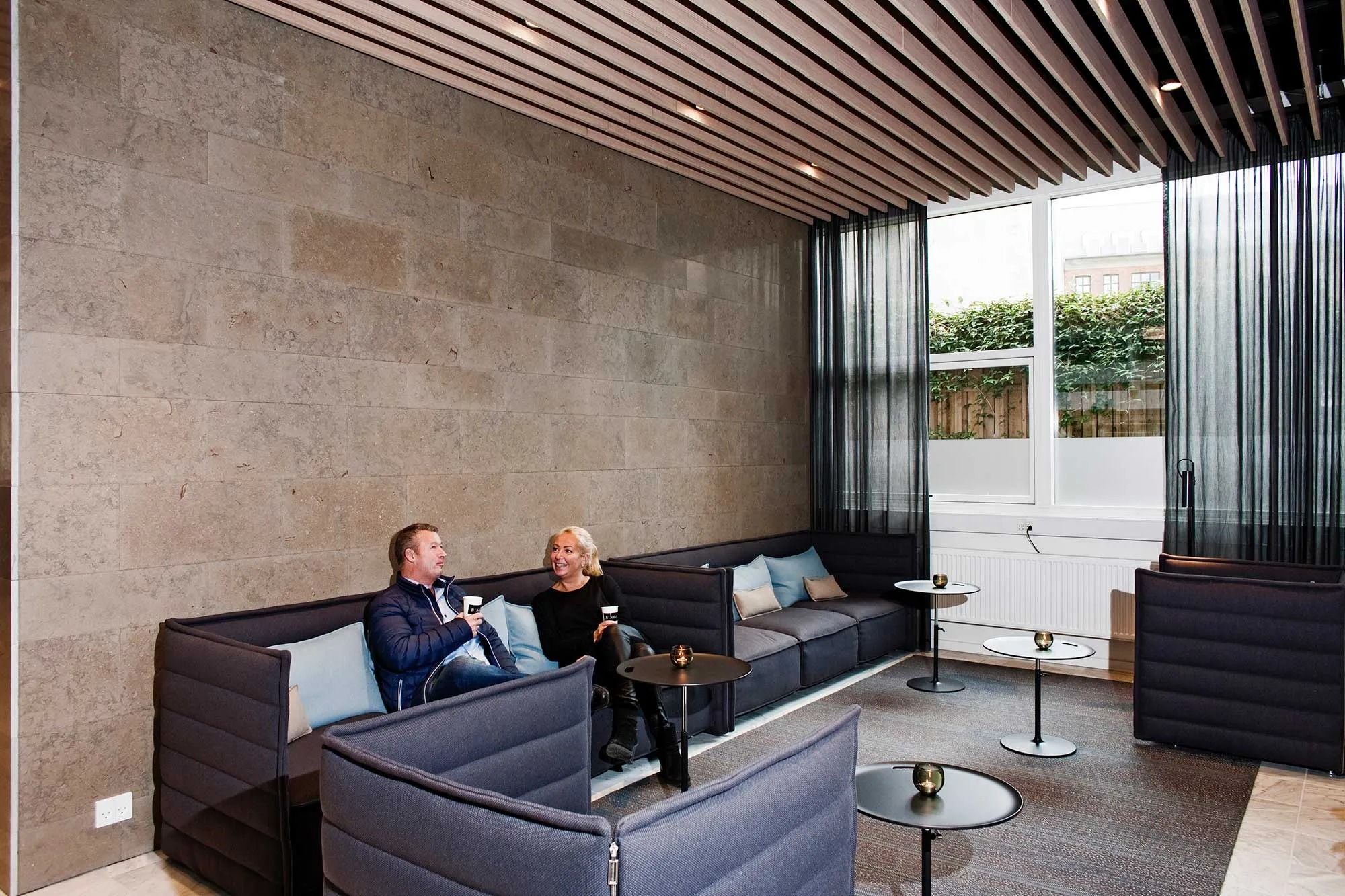 To mennesker sidder og nyder kaffe på en sofa i et moderne kontor med møbler og træpaneler