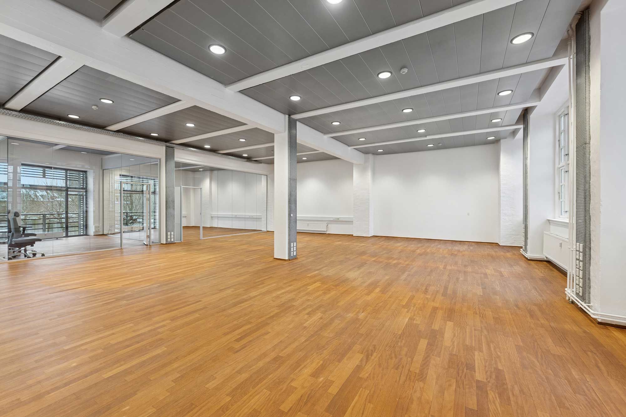 En stor, tom kontorlokal med høje lofter og glasdøre i hvide vægge og massivt trægulv samt hængende loftslampe med flere runde lysekroner