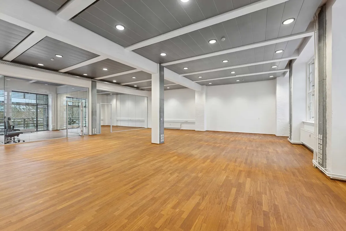 En stor, tom kontorlokal med høje lofter og glasdøre i hvide vægge og massivt trægulv samt hængende loftslampe med flere runde lysekroner