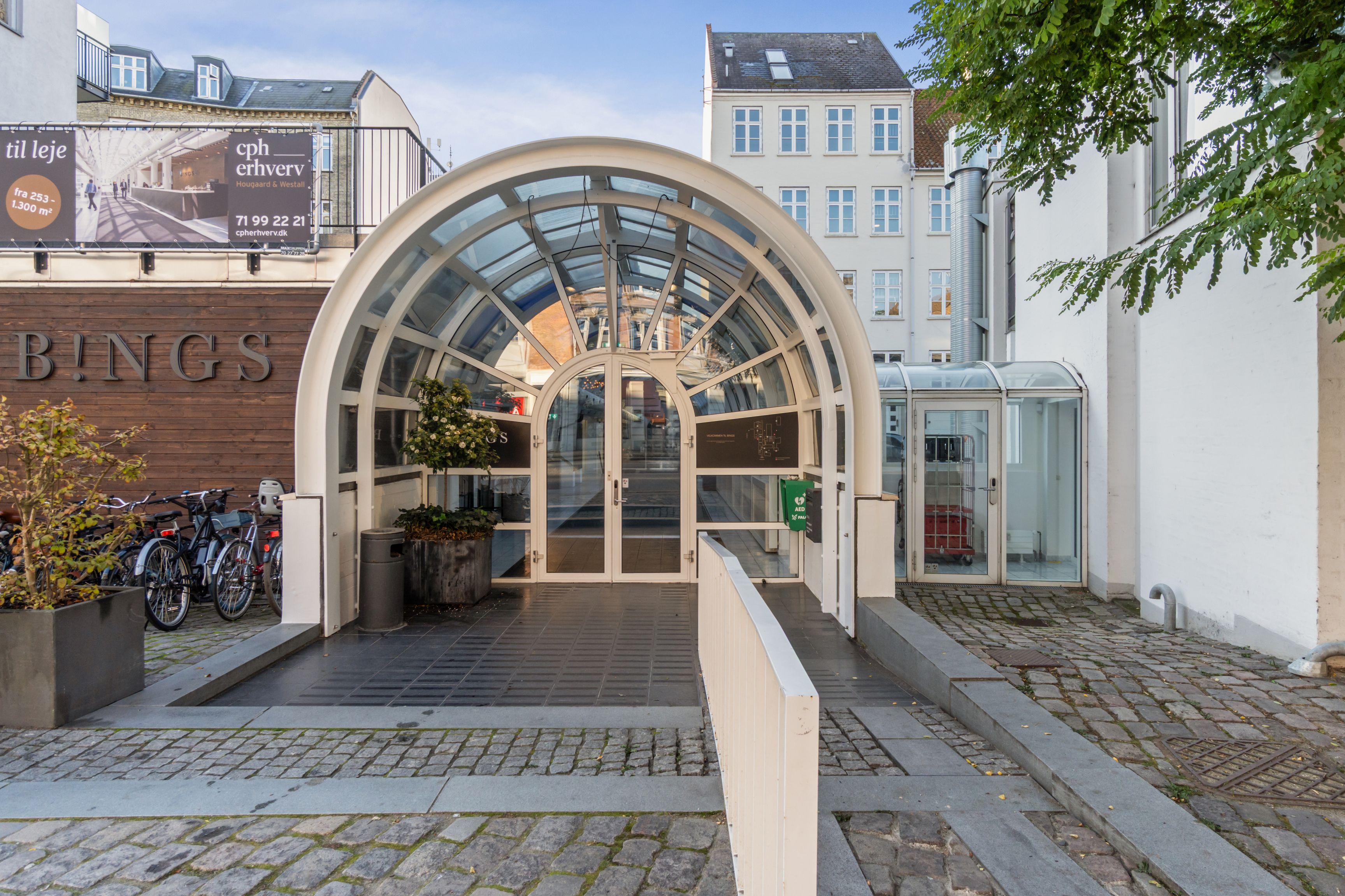 Forretningskontor i bygning med glaskuppel og vinduer, facade med teksten Bings, gulv med røde mursten, hvid trappe og kunstplanter i potter, og cykler parkeret udenfor