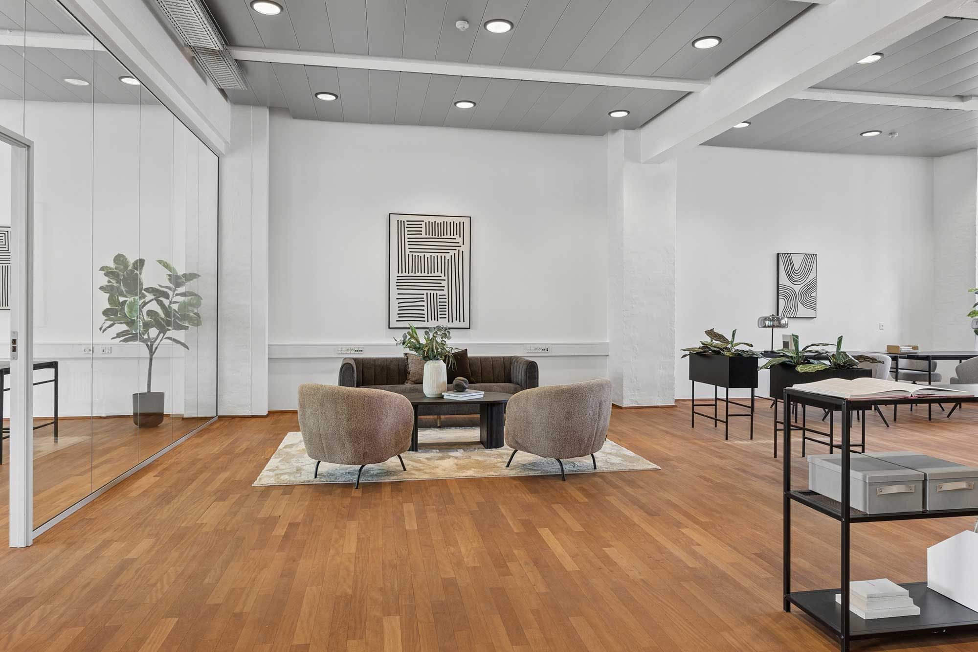Minimalistisk kontor med højt loft og hvide vægge, lyse trægulve og moderne møbler, inklusiv to sædestole og et sofa. Sorte plantepoter og en stor plante, og et skrivebord med et åbent bog