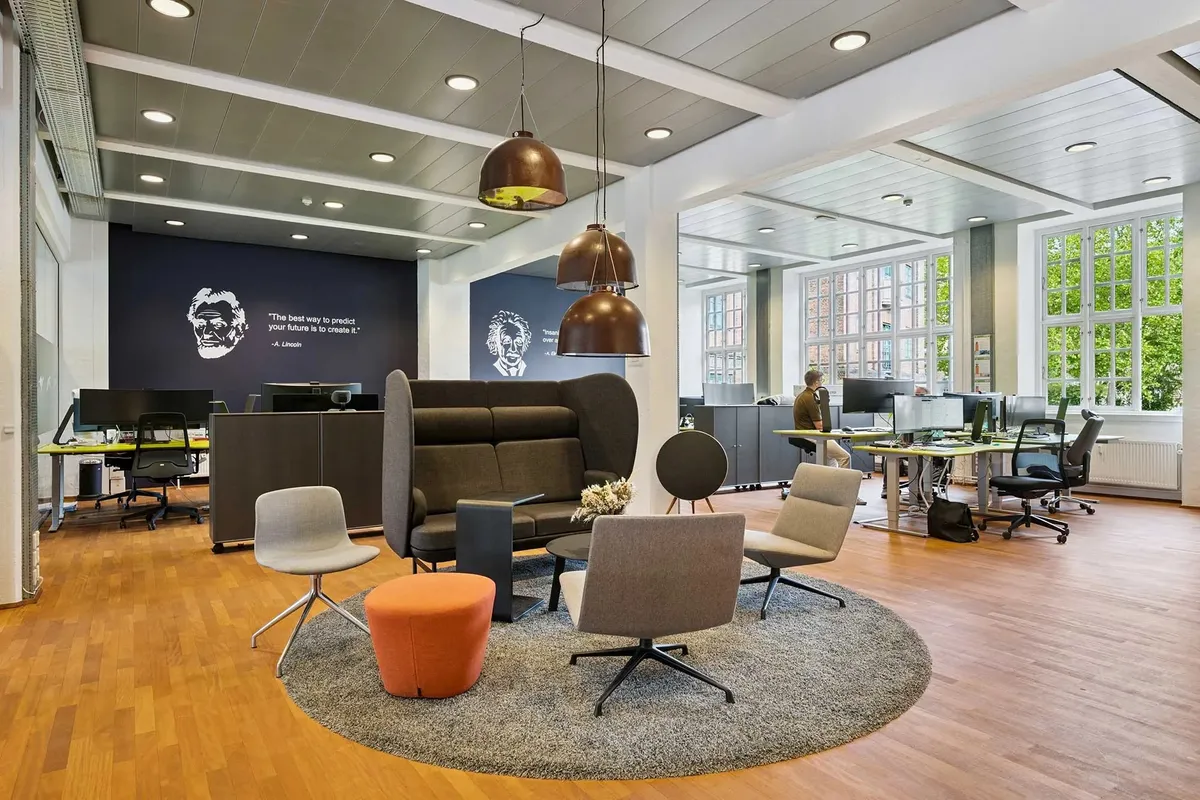 Moderne kontor med lyse loftsbelægninger, rundt bord med møbler og lyse lamper og mange skriveborde og computere