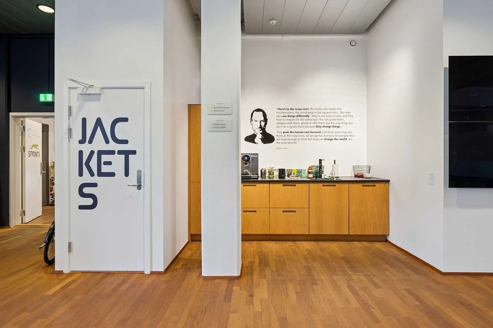 Inde i et kontorrum med åbne døre og gulvbelægning af træ, tekst "Jackets" og portræt af Steve Jobs, der er fundet på væggen