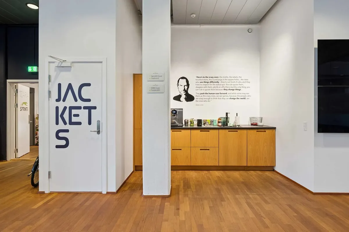 Inde i et kontorrum med åbne døre og gulvbelægning af træ, tekst "Jackets" og portræt af Steve Jobs, der er fundet på væggen