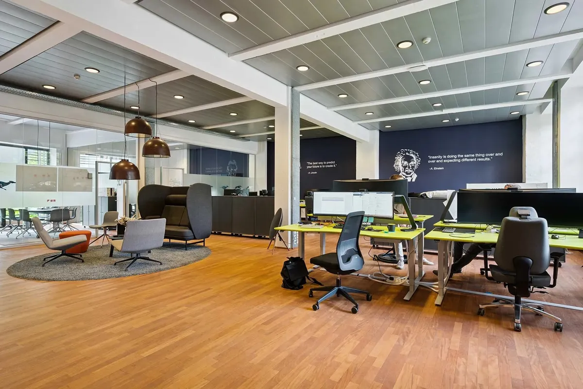 Lyst og moderne kontor med trægulve, skriveborde, skrivebordsstole og loftslys. Store vinduer, stolpe og gulvvægge. En