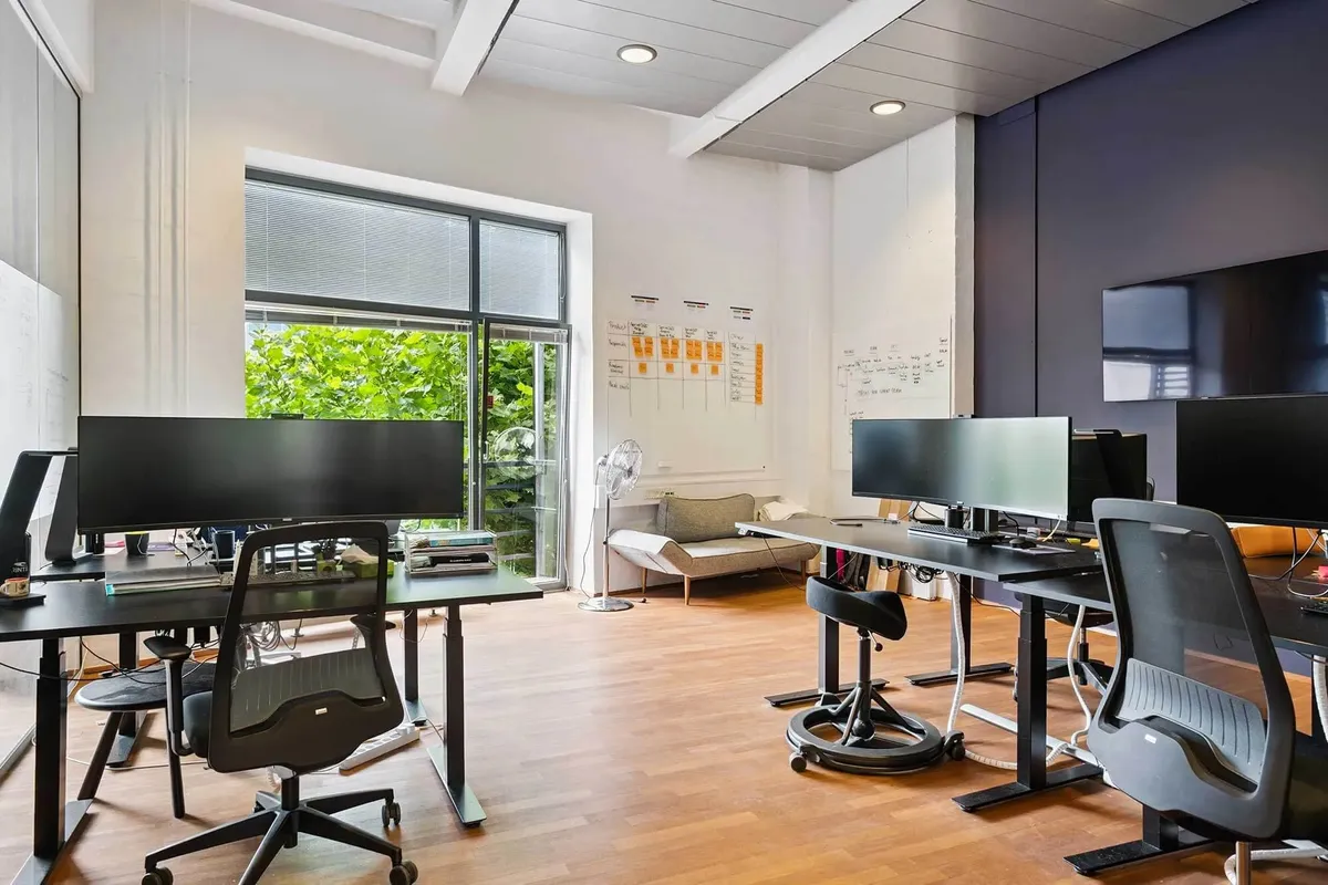 Kontor med skriveborde og ergonomiske sædestole, moderne monitore og skrivebordsventilator og sofa i baggrunden