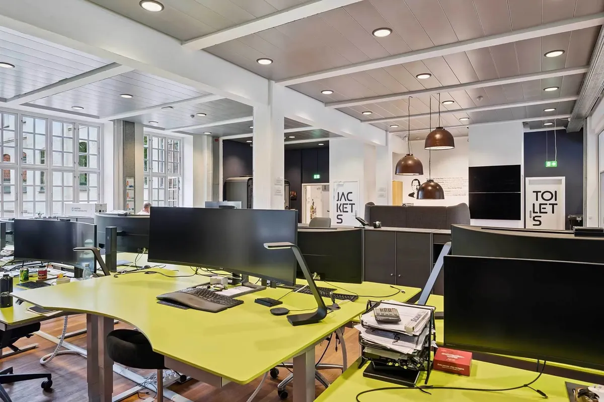 Et stort kontor med lysegulle skriveborde og lysebrune gulve. To skriveborde med to skærme og en stol. En person i