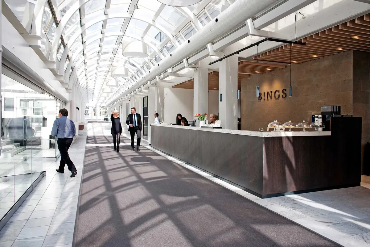 Folk går gennem en hall med glas og marmor. En reception er placeret med borde og en plante i hvid vase.