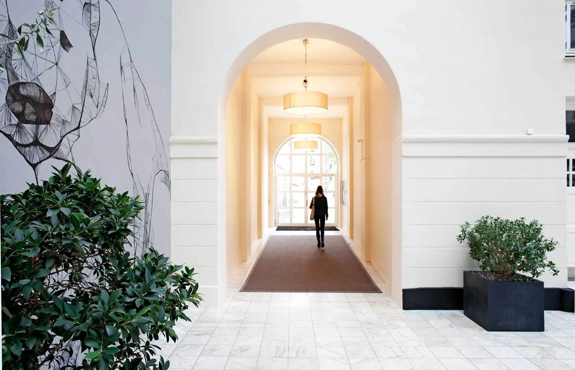 En kvinde går gennem en lys hall med hvide vægge og to hængelampe, en plante i sort potte og en panda tegner på væggen