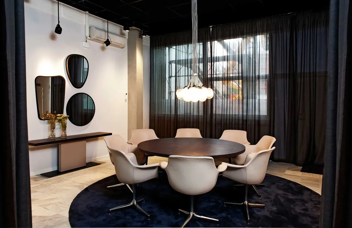 En rund bord med flere hvide stole omkring i et moderne kontor. Et rundt, blåt tæppe ligger under bordet. Tre spejle er