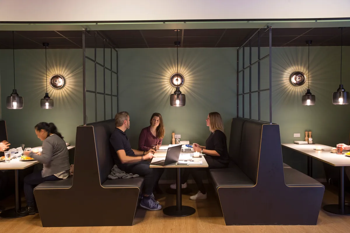 Fire personer sidder ved borde og bruger laptopper og skriveborde i moderne kontor med loftsbøjer og elegante lamper og lyse vægge